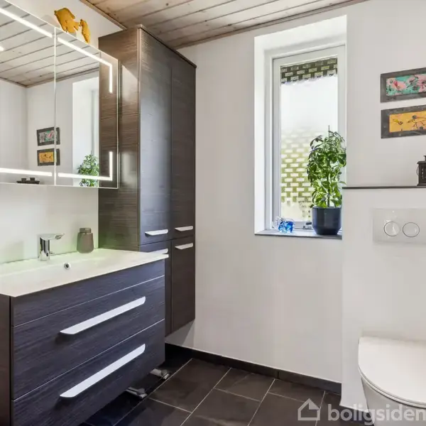 Et moderne badeværelse med mørke skabe og spejl over en vask. Et vindue med en plante ved siden af toilettet på gulv med sorte fliser.