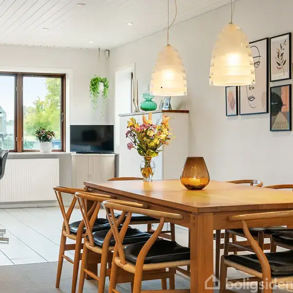 Spisebord med blomster i vase placeret under to hængende lamper; omgivet af stole. Vægbilleder og lys indretning i opholdsrum med vindue og fjernsyn i baggrunden.