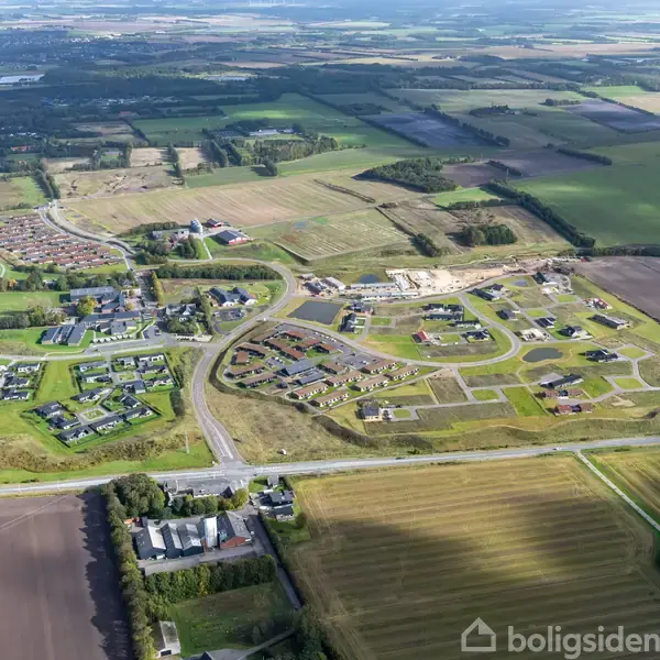 Et parcelhuskvarter med spredte huse ses fra luften, omgivet af marker og skove i et landligt område. Vejnettet forbinder de forskellige bebyggelser, mens åbne marker breder sig omkring.