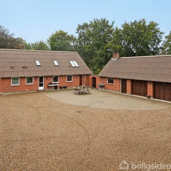 En rød murstensbygning med skråt tag står stille. Ved siden af er en garagebygning. Grusvej omgiver bygningerne, og træer i baggrunden skaber en grøn omgivelse.