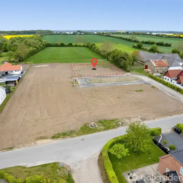 Grusvej markerer en ubebygget grund omgivet af huse og marker. Marker er gyldne og grønne under en klar himmel. Røde hustage ses i nærheden, med fokus på sort plet på jordgrunden.