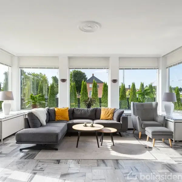 Stor, lys stue med en grå hjørnesofa og lænestol, ved siden af et rundt sofabord. Store vinduer slipper meget lys ind. Grøn have ses udenfor. Ingen tekst.