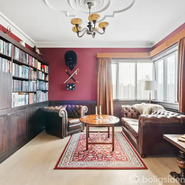 Stue med mørkerøde vægge og brune lædersofaer. Et sofabord står på et mønstret tæppe. Reoler fyldt med bøger dominerer rummet. Lysekrone hænger i loftet.