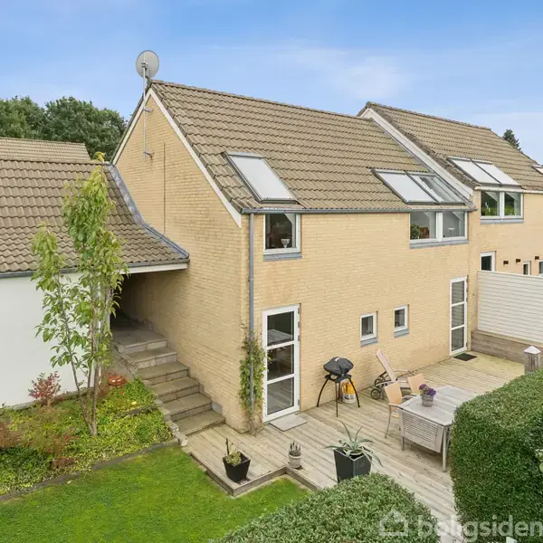 En beige murstensvilla med tagvinduer og terrasse i en velholdt haveindretning. Flere trappetrin forbinder terrassen med det grønne græsareal, omgivet af hække.