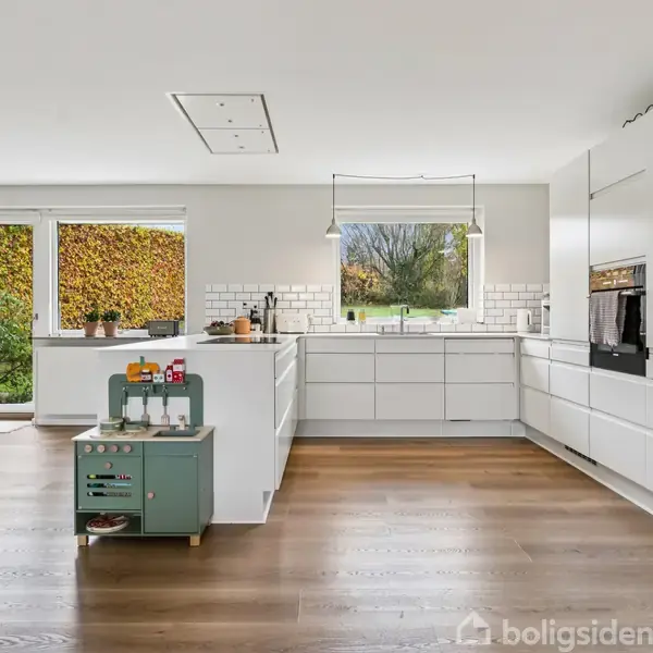 Køkken med hvide skabe og integrerede apparater står stille i et rummeligt, lyst rum. Store vinduer til venstre og centralt giver udsigt til træer og grønne omgivelser.