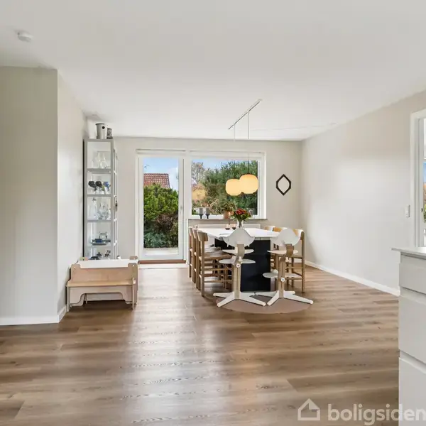 Spisebord står centralt med seks stole placeret omkring det. Store vinduer i baggrunden giver udsigt til en have. Rummet har trægulve og lyse vægge, skabe til venstre.