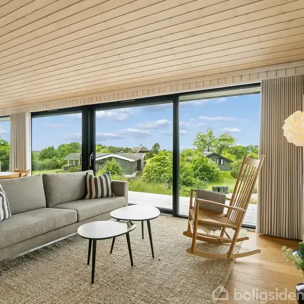 Grå sofa står foran gulv-til-loft-vinduer med udsigt over grønt landskab; en gyngestol ved siden af, indendørs planter og moderne lamper skaber en hyggelig stueatmosfære.