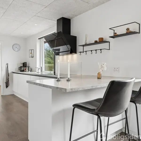 Køkkenbar med to sorte barstole, under en grå bordplade, omgivet af åbne hylder og vindue til en have. Vægge er hvide, og gulvet er af træ.