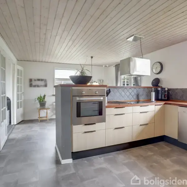 Et moderne køkken med lyse skabslåger og integreret ovn, står ud i et lyst rum. Køkkenet har grå gulvfliser, træloft, og en kaffemaskine ved siden af et vægmonteret ur.