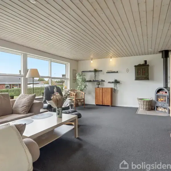 Stue med beige hjørnesofa og sofabord, foran store vinduer, der giver naturligt lys. Der er en brændeovn i hjørnet og hylder med planter på væggen.