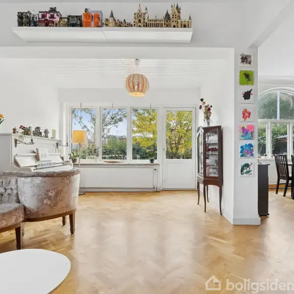 Et stueområde med en elegant sofa og klaver i et lyst rum. Store vinduer viser grønne træer udenfor, mens en spisestue med sorte stole ses til højre. Bøger fylder bogreolen til venstre. Kunst dækker væggen.