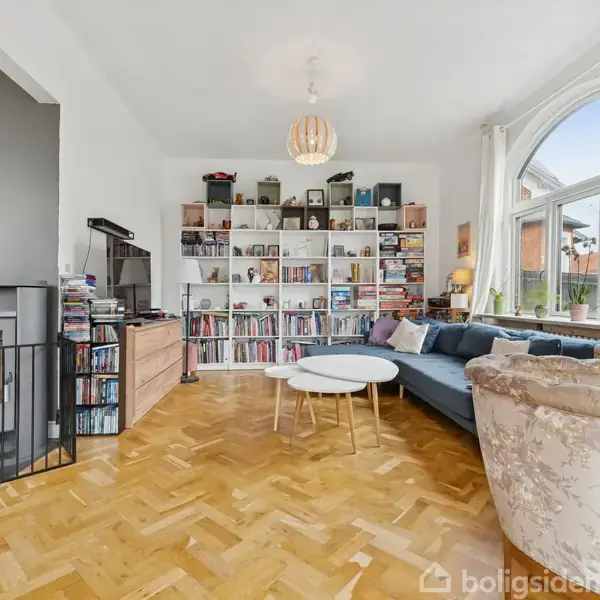 En stue med en reol fyldt med bøger og dekorationer, en blå sofa ved siden af et rundt sofabord. Rummet har store vinduer og en pejs i venstre hjørne.