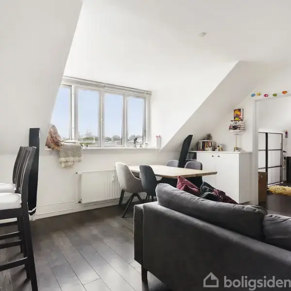 Stue med sofa og spisebord, lyse vægge og loft, skråvægge, stor vindueskarm, farverige pyntelamper. Mørkt trægulv og lysindfald skaber en lys og rummelig atmosfære. Indgangen ses i baggrunden.