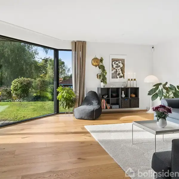 En stue med en grå sofa ved siden af et lille bord og en sort lænestol. Store vinduer giver udsigt til en grøn have. En reol og planter dekorerer rummet.