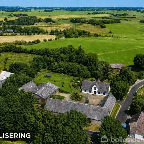 En landlig ejendom set ovenfra med flere bygninger omgivet af frodige marker og skov, ligger ved en snoet vej. Tekst: "VISUALISERING".