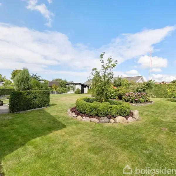 Grønne græsplæner omkranset af hække og buske med blomster i midten. En flagstang står i baggrunden. Huse og en terrasse ses omkring haven, under en blå himmel med skyer.