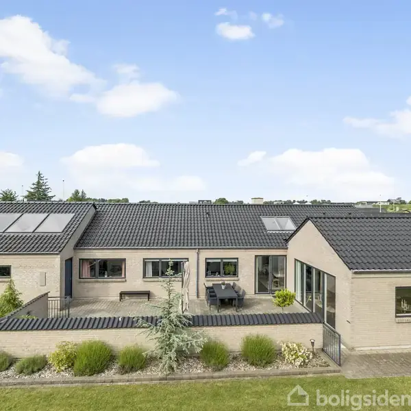 En moderne, flad tagstensvilla med solpaneler og store vinduer står i et velplejet haveområde med grønne buske og en terrasse. Himlen er blå med få skyer.