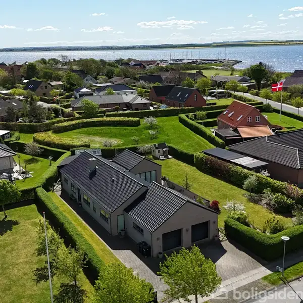 Hus med sort tag i forreste have, omgivet af velplejet græsplæne og hække, i en forstad i nærheden af vand. Røde huse og en dansk flag i baggrunden.