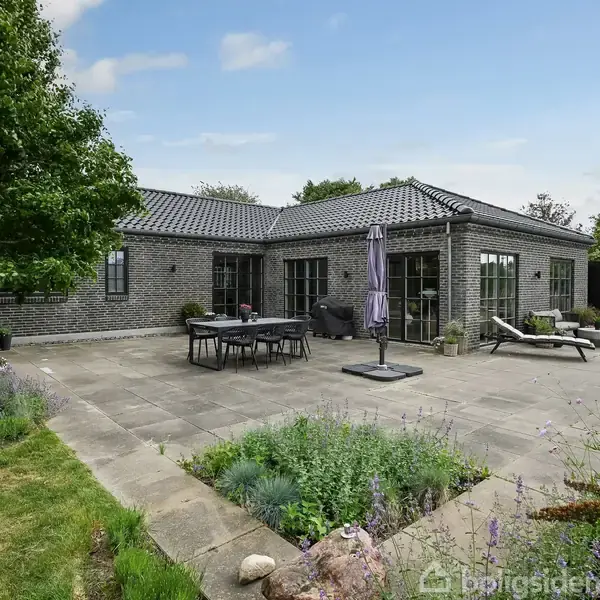 Et moderne hus med gråt murstensfacade står roligt. En stor fliseterrasse med havebord, stole og liggestole omringes af grønne buske og planter.