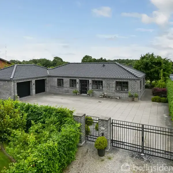 En ensidet murstensvilla står stille med sorte tagsten, omkranset af frodig hæk og have. Flisebelagt indkørsel og dobbelte garageporte ses foran huset.