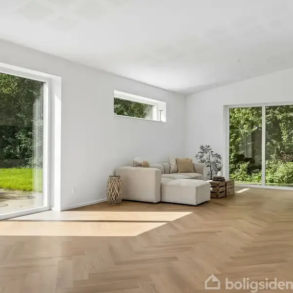 Stue med hvid væg og træparket, en lys sofa i hjørnet ved store vinduer. Udsigt til have med træer gennem glasset. Sofa flankeret af sidebord og plante.