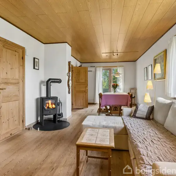 Brændeovn opvarmer hjørne af stue med træmøbler og komfortabel sofa. Trægulv og -loft skaber hyggelig atmosfære. Store vinduer giver udsigt til naturen udenfor. Bord med lilla dug i baggrunden.