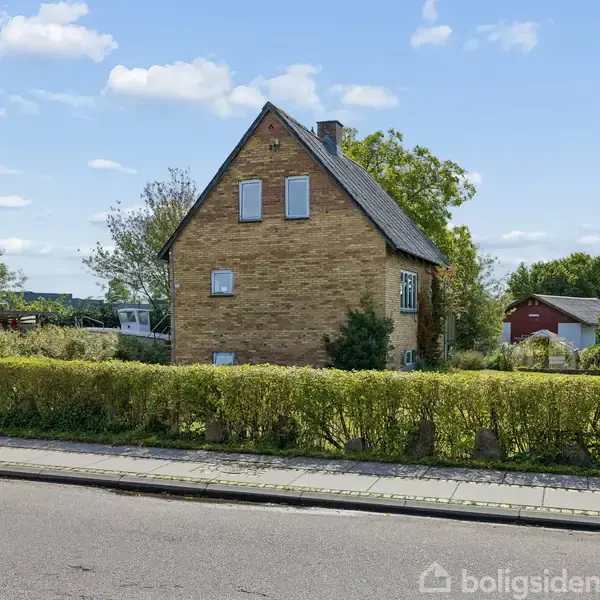 Et gult murstenshus står stille omkranset af en lav hæk i et villaområde med træer og en blå himmel i baggrunden.