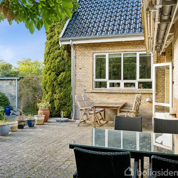 Terrasse med glasbord og stole, omgivet af grønne planter og blomsterpotter. Bagved ligger et murstenshus med et vindue og en åben dør. Solrig og velholdt haveatmosfære.