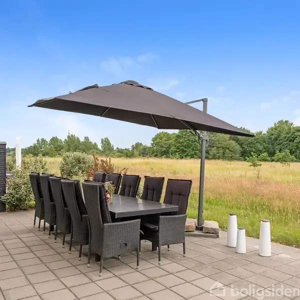 Et sort spisebord med ni stole står under en stor, sort parasol på en terrasse med fliser. I baggrunden er en åben mark og træer under en blå himmel.