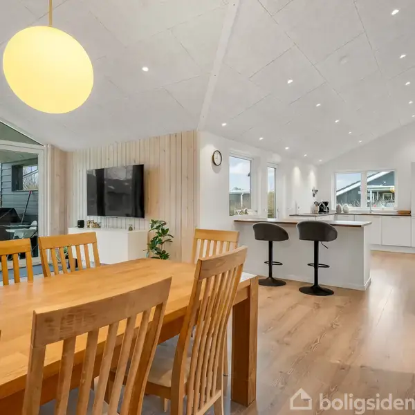 Spisebord med træstole står i en moderne stue-køkken-alrum med hvide vægge og trægulv. I baggrunden er et køkken med barstole, og store vinduer viser en have med gyngestativ.