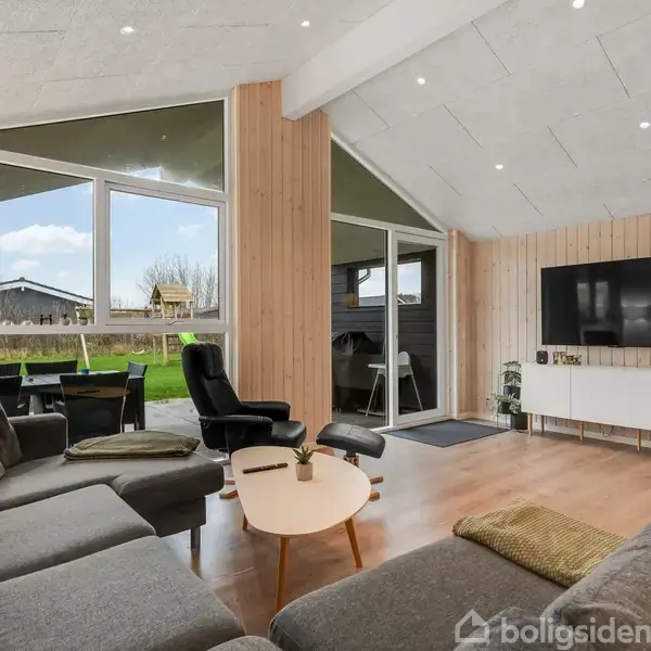 En stue med grå sofa og sort lænestol ved et rundt bord, set mod en stor fladskærm på væggen. Store vinduer viser en have med græs og himmel udenfor.