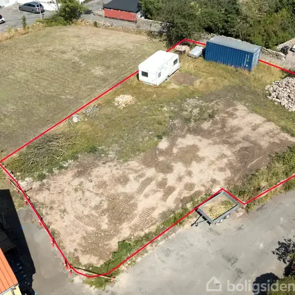 Grunden er indhegnet med en rød linje, hvor der står en campingvogn og en blå container. Omgivelserne inkluderer græs, stenbunker og nærliggende bygninger.