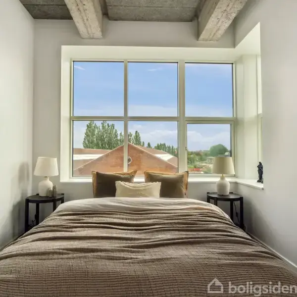 Seng dækket med brunt sengetøj står foran stort vindue med udsigt til træer og blå himmel. Natborde med lamper står på hver side. En trappestige pusterum hænger til højre.