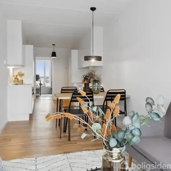 Stue med moderne møblering, hvor en sofa står til højre. Et spisebord med stole står centralt. Der er en køkkenbaggrund med lyse skabe. Kunst på væggen til højre.