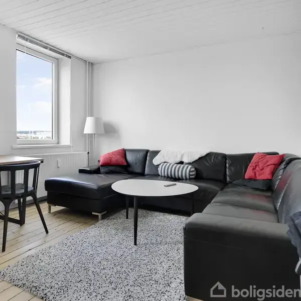 Sort hjørnesofa med farverige puder står på lyse trægulve ved siden af rundt, hvidt sofabord. Spisebord med stole og lampe tæt på vindue og hvid væg til venstre.