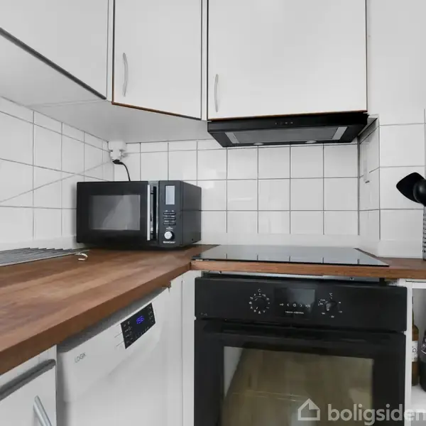 Et kompakt køkken med en indbygget ovn og kogeplade. På bordpladen står en mikroovn ved siden af et bestikholder med køkkenredskaber. Hvide skabe hænger over en flisebeklædt væg.