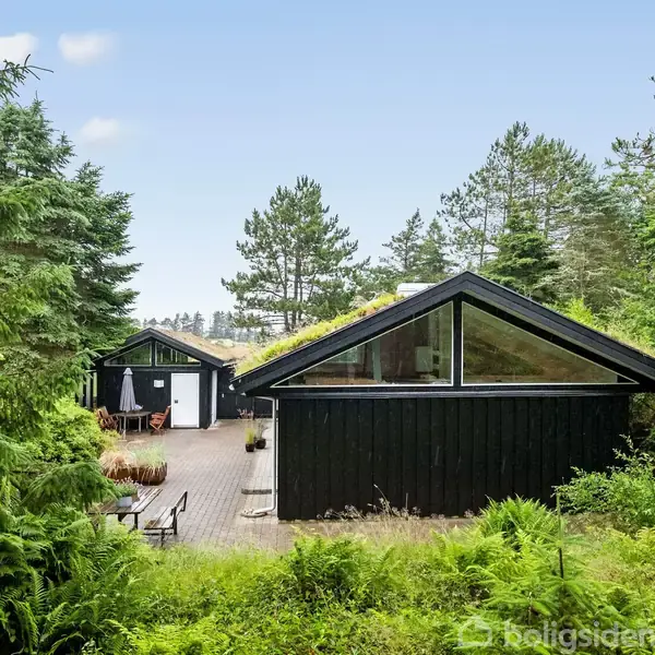 Sort træhus med store glaspartier står stille omgivet af tætte grønne træer og buske. Terrassen har havemøbler og en parasol, hvilket skaber en fredelig, naturlig atmosfære.