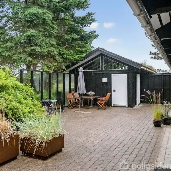 En flisebelagt gårdhave med planter og et bord med stole ved siden af et sort skur. Omkranset af træer og buske, skaber en stille, naturlig atmosfære.