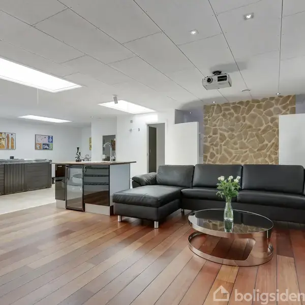 Sort lædersofa med glasbord foran i et moderne opholdsrum. Baggrunden indeholder køkken med mørke træelementer og moderne kunstværker. Loftet har indbyggede lys og projektor. Plankegulv skaber hyggelig atmosfære.