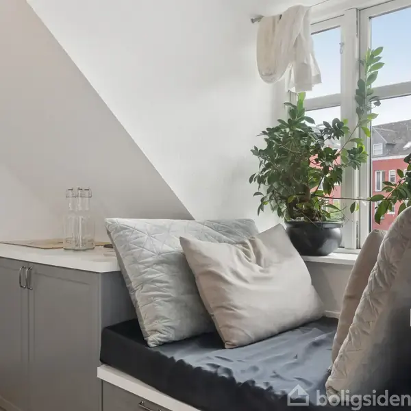 En hyggekrog med grå skabssektioner fungerer som siddeplads ved et vindue. Puder ligger på bænken, og der er grønne planter på vindueskarmen.