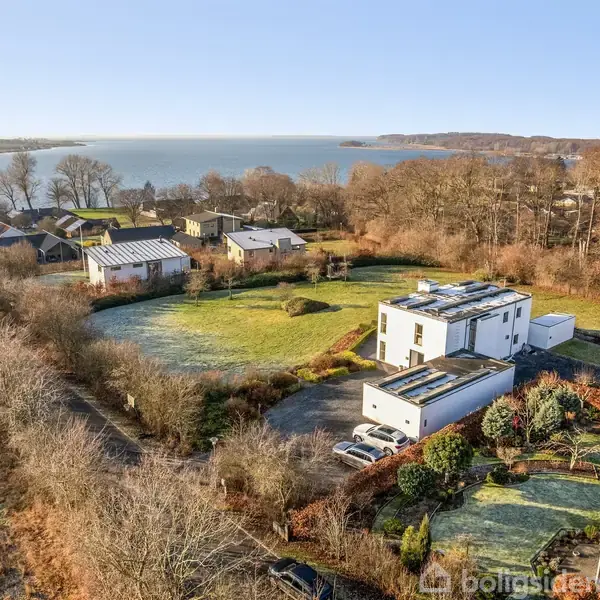Et hvidt moderne hus ligger i et åbent landskab ved en sø med andre boliger og skov i baggrunden. Klar blå himmel og solskinslys skaber et fredeligt miljø.