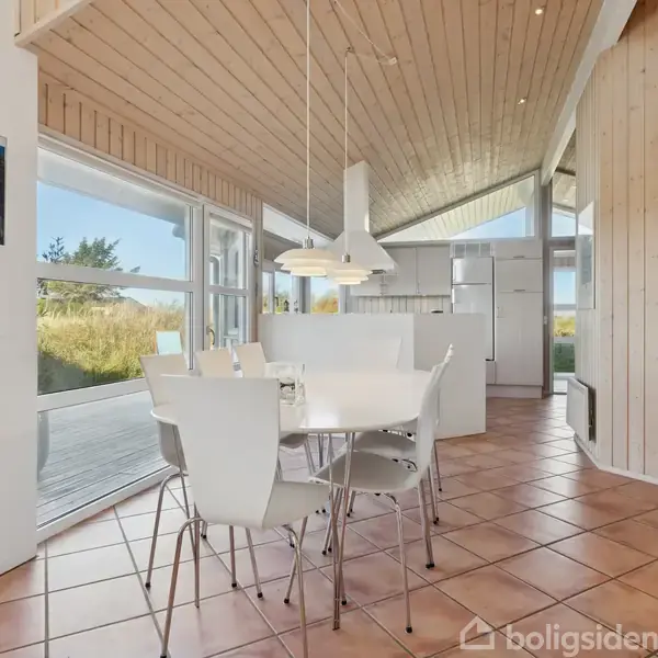 Et spisebord med seks hvide stole står i et rum med højt til loftet. Store vinduer til venstre giver udsigt til en terrasse og natur udenfor.