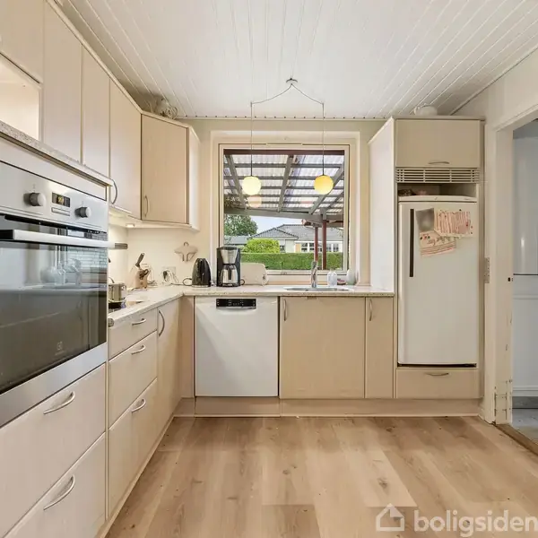 En gul murstensvilla med tegltag ligger blandt grønne haver. Der er en overdækket terrasse og tæt beplantning omkring huset. Omkringliggende huse ses i en forstadslignende bebyggelse.