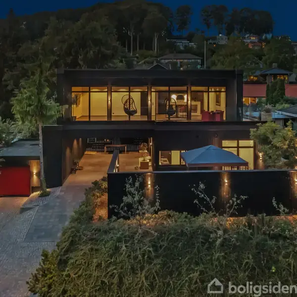 Moderne hus oplyst om natten med stor glasfacade, hvor man kan se ind i stuen. Det er beliggende i et beboelsesområde omgivet af træer og andre huse.
