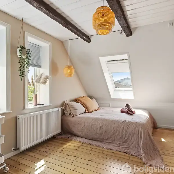 Seng under loftsbjælker i solbelyst soveværelse med trægulv; omgivet af vinduer med planter og et lille bord fyldt med toiletartikler. En varm og hyggelig atmosfære dominerer rummet.