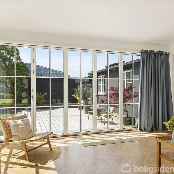 Stue med store vinduer slipper lys ind. Stol og bord med plante i forgrunden. Terrasse og have udenfor. Blå gardiner til højre. Læsehjørne med lampe til højre.