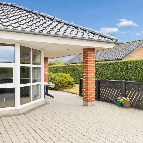 Terrasse med flisebelægning, store vinduespartier i muren med røde mursten, omgivende hæk og træhegn med havekrukker; under blå himmel med spredte skyer.