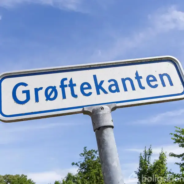 Et vejskilt med teksten "Grøftekanten" står på en metalstang. Skiltet ses mod en baggrund af blå himmel og trætoppe.
