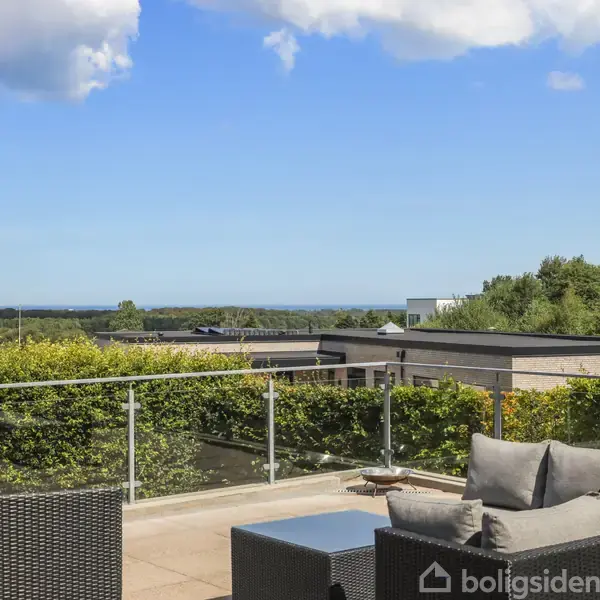 Udendørs terrasse med grå sofa og glasrækværk. Sofaen står stille. I baggrunden er der udsigt over grønne træer og blå himmel med enkelte skyer.