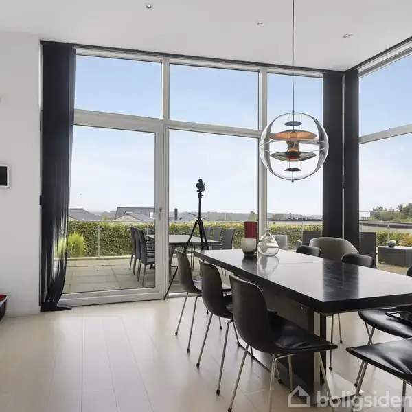 Spisebord med otte stole står i et moderne rum med store vinduer. En farverig lampe hænger over bordet. Udsigt til terrasse og have gennem glasdør.